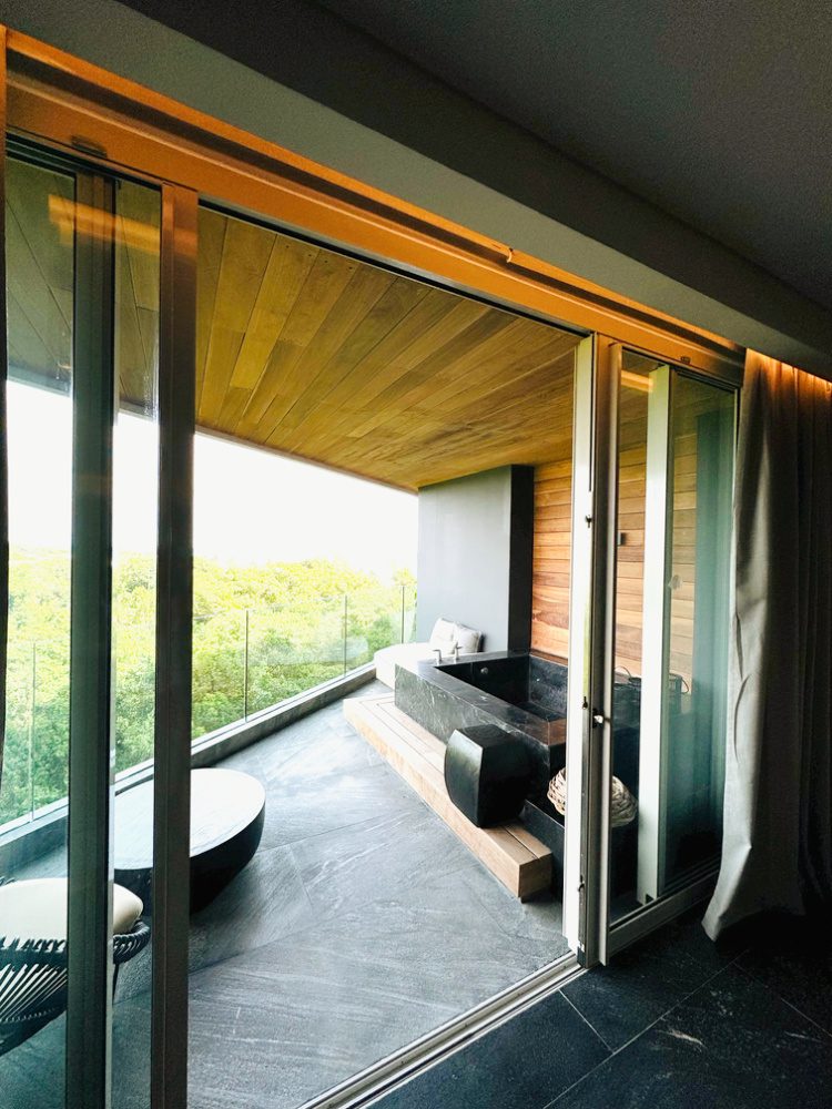 Photo of large double sliding doors leading onto a spacious black granite balcony with a two-person soaking tub and comfortable outdoor seating. The balcony is surrounded by teak wood and a glass railing that looks out onto a lush wooded area and the ocean.