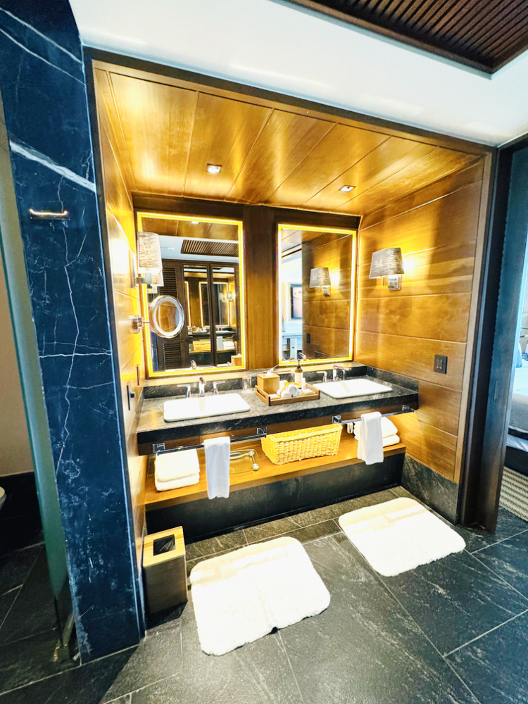 Photo of brightly lit bath counter with double sinks and mirrors, surrounded by modern teak accents and black granite in the guest suite at all-inclusive Impression Moxché in Playa Del Carmen, Mexico.